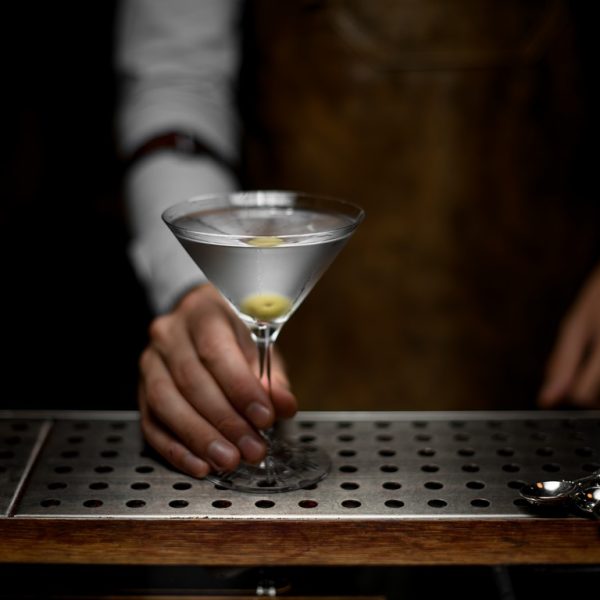 Male bartender holds cocktail glass with martini on metal bar counter